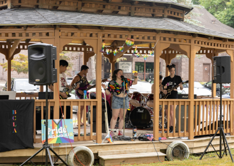 A band with guitar, bass, drums and a female vocalist performs in a gazebo.