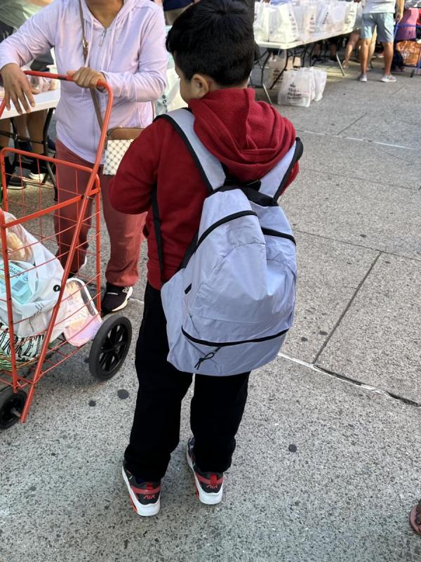 A boy shows off his new blue backpack as families in the background pick up donated school supplies.